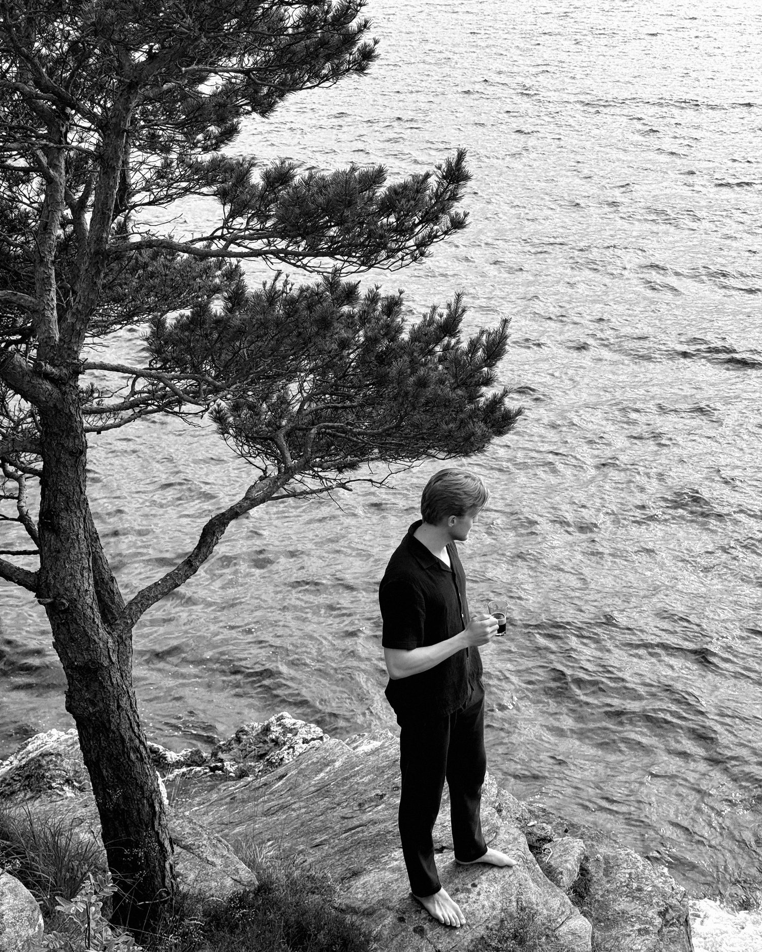 Tarjei står barbeint på svaberget med en kaffekopp og ser utover sjøen, en furu over seg.